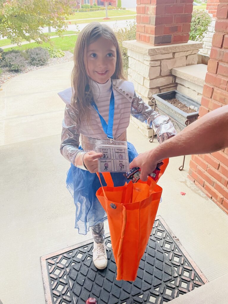 Young girl dressed in costume holds out her treat bag at someone's door. She is wearing a lanyard with picture symbols for trick or treat, thank you, happy Halloween and boo.  