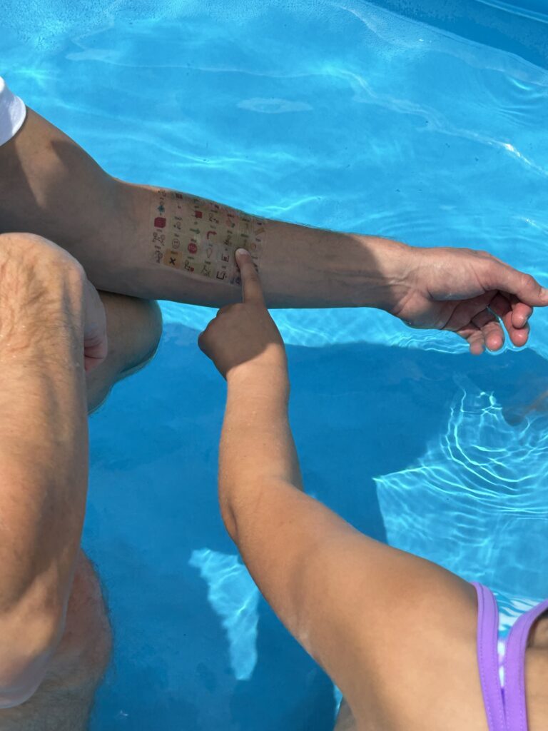 Child in the pool pointing to a communication icon on a swim tattoo on her father's arm.