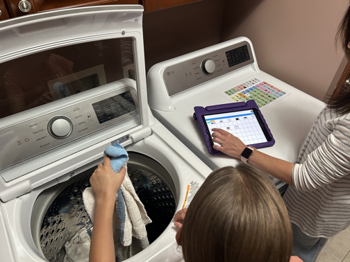 Mother modeling a word on a speech generating device while her child is putting laundry into the washing machine
